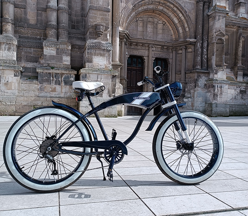 Beach cruiser Mecabecane bleu marine et blanc, vu de profil dans un paysage urbain